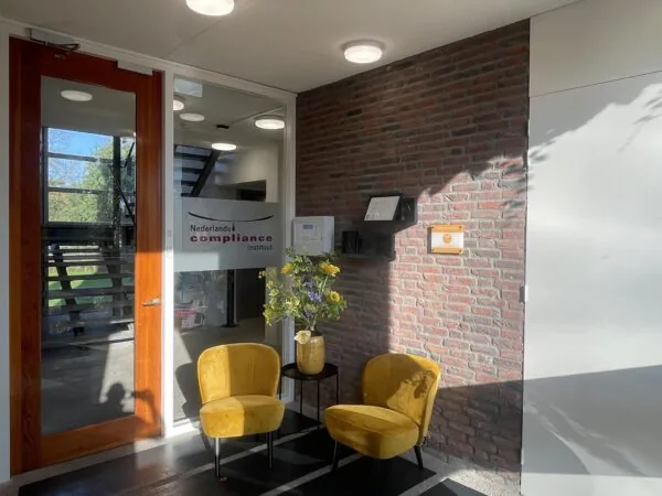 Inkomhal van het Nederlands Compliance Instituut met twee gele stoelen en een bloemenvaas tegen een bakstenen muur.